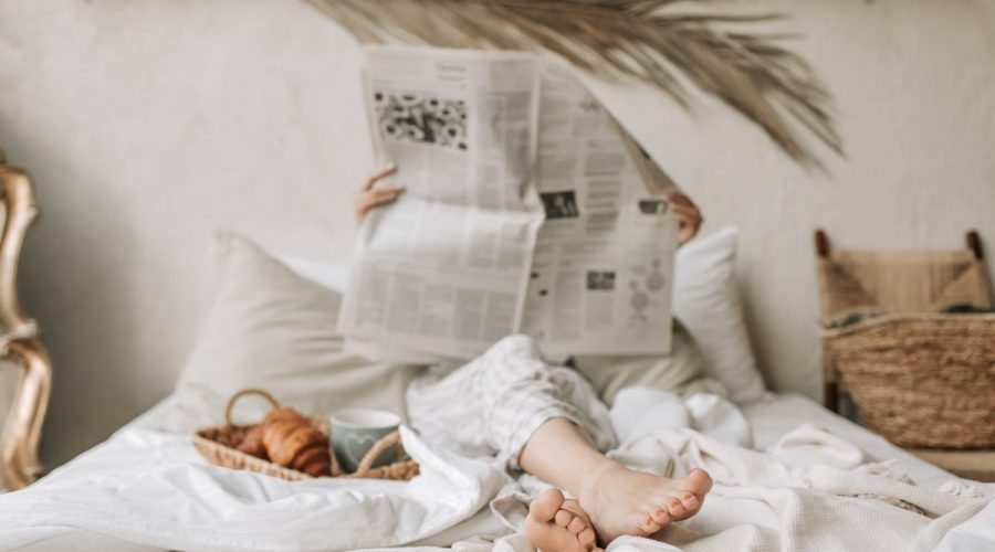 Person relaxing in bed with a newspaper, coffee, and croissant for a calm morning vibe.